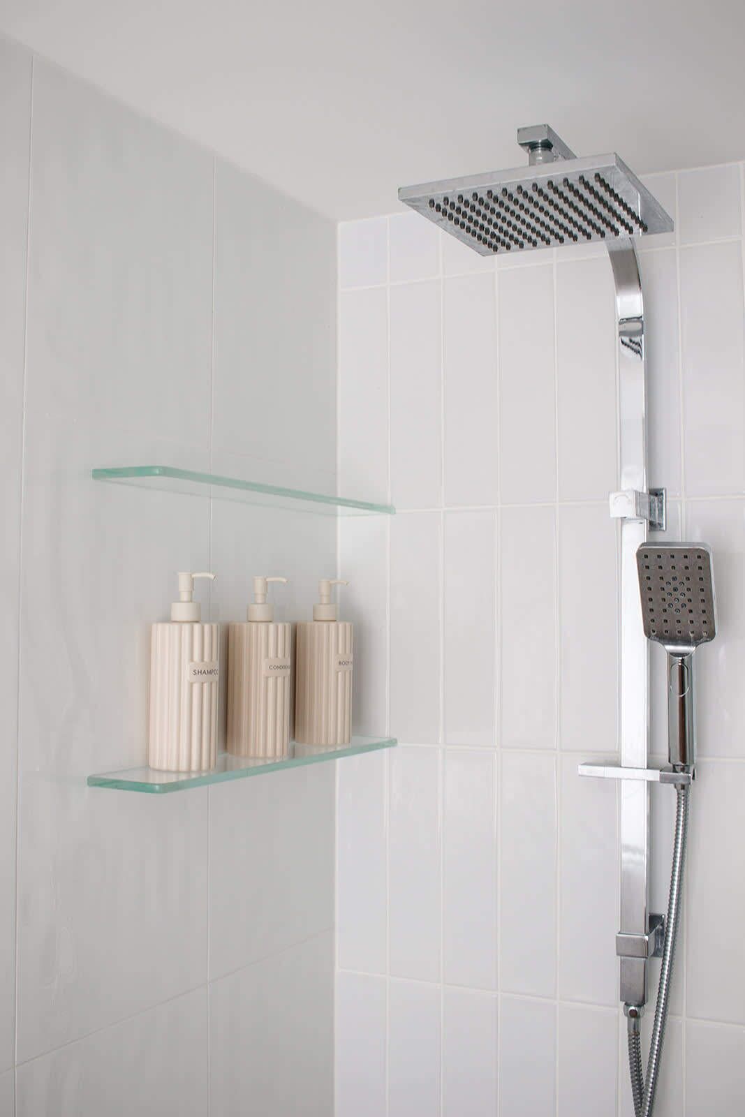 Eine Dusche mit Duschkopf, Handbrause und Ablagefläche aus Glas mit drei Flaschen.
