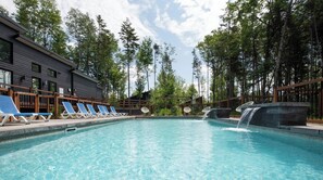 A swimming pool with lounge chairs and a waterfall feature, surrounded by trees and a building with a dark facade.