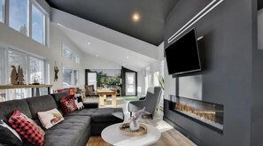 A modern living room with a fireplace, a flat-screen TV, and a dining area visible through an open door.