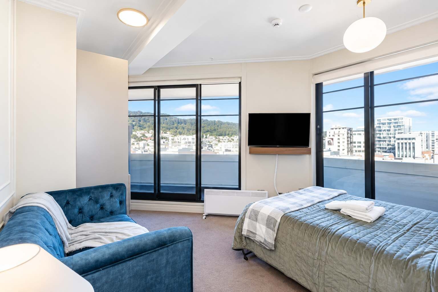 Quarto de hotel moderno com uma cama grande, uma poltrona azul e uma televisão fixada na parede.