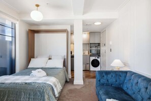 A bedroom with a bed, a blue sofa, a washing machine, and a kitchenette.