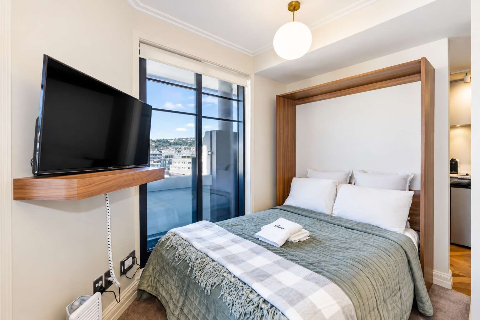 Quarto de hotel com uma cama, televisão presa a uma prateleira de madeira, uma janela grande e vista para a cidade.