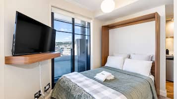 Quarto de hotel com uma cama, televisão presa a uma prateleira de madeira, uma janela grande e vista para a cidade.