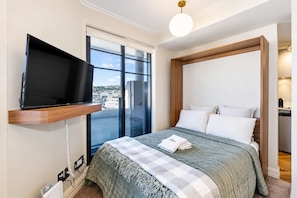 A hotel room with a bed, a television mounted on a wooden shelf, a large window, and a view of the city. - City Lookout | Te Aro Studio + Private Balcony (Wellington)