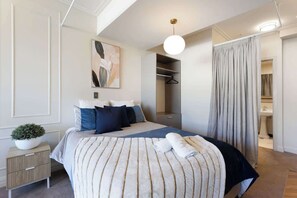 Ein modernes Schlafzimmer mit einem großen Bett, einer Zimmerpflanze, einem Kleiderschrank und einem durchgehenden Türrahmen einsehbaren Badezimmer.