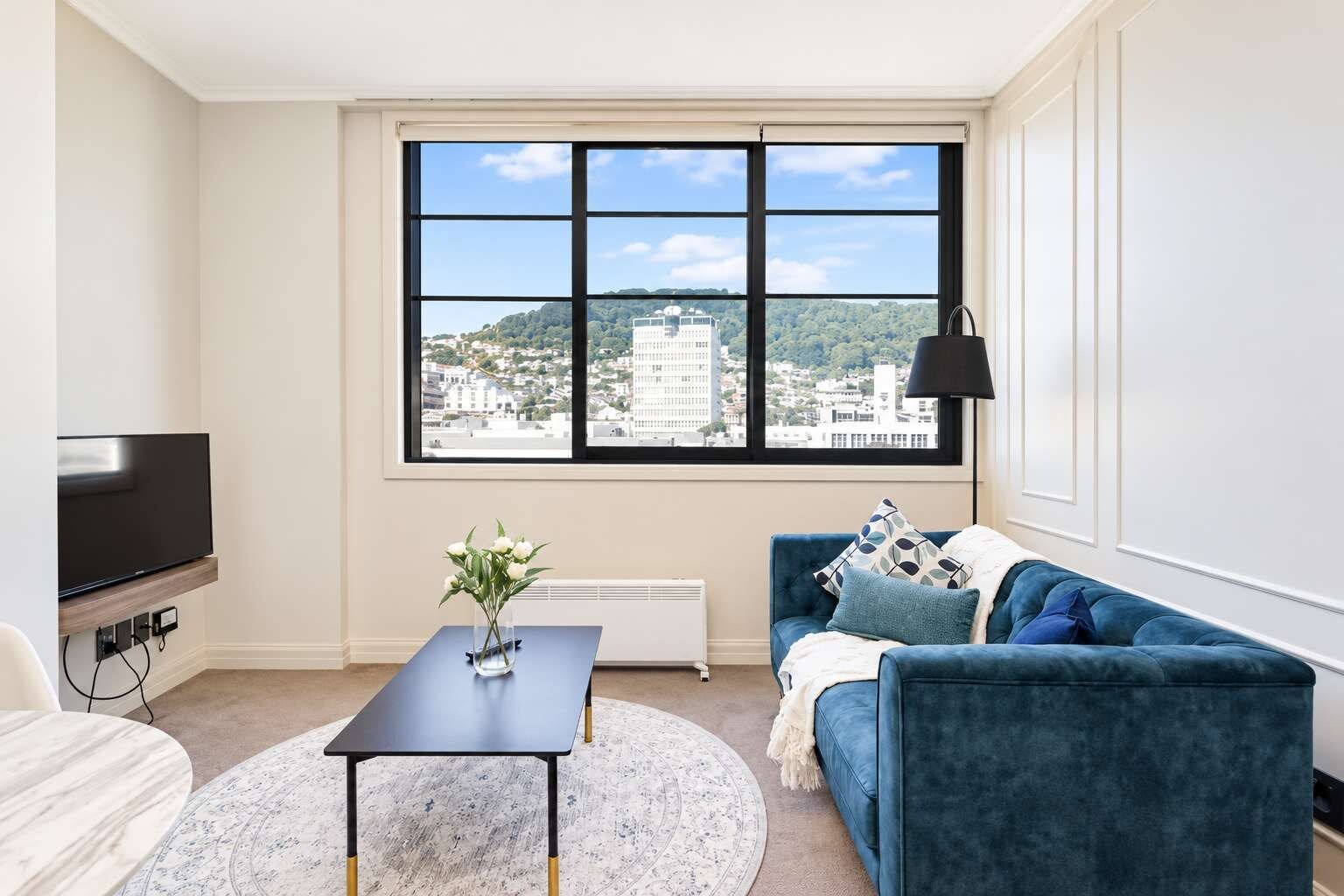 A modern living room with a large window, a flat-screen TV, a blue sofa, and a round coffee table.
