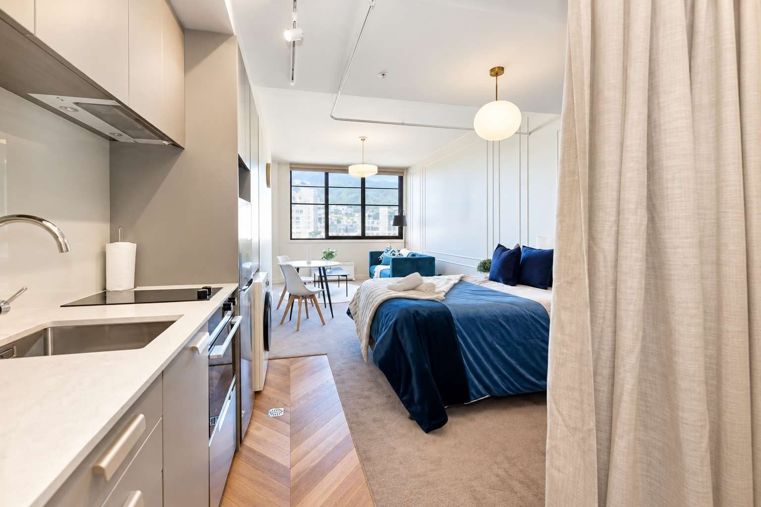 A modern hotel room with a kitchenette, a bed with blue bedding, a desk, and a large window offering a city view.