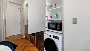 Een kleine wasruimte met een wasmachine, een magnetron en opslagplanken.