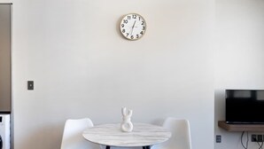 A modern dining area with a round table and two chairs, a wall-mounted clock, and a television on a stand. - Cityview Studio + Kitchen | Wellington CBD (Wellington)