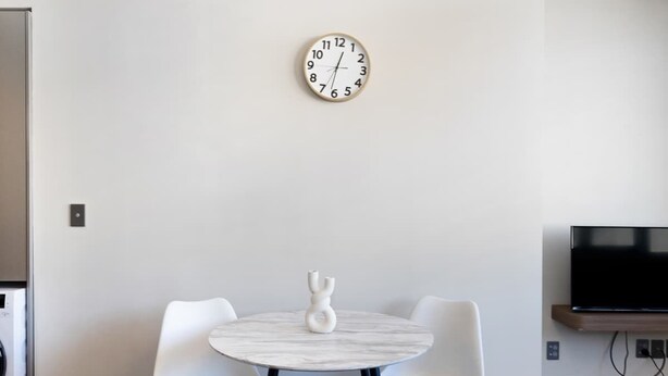 A modern dining area with a round table and two chairs, a wall-mounted clock, and a television on a stand.