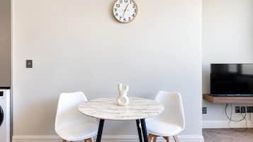 A modern living room with a round marble table, two white chairs, a wall clock, and a flat-screen TV.