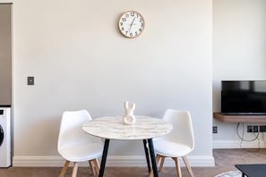A modern living room with a round marble table, two white chairs, a wall clock, and a flat-screen TV. - Cityview Studio + Kitchen | Wellington CBD (Wellington)