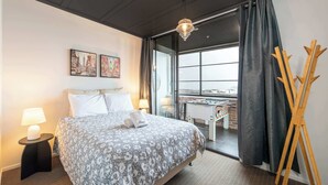 A hotel room with a bed, a side table with a lamp, artwork on the wall, and a view through a large window.