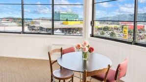 A room with a round table, chairs, a vase with flowers, and large windows offering a view of buildings and a parking lot.