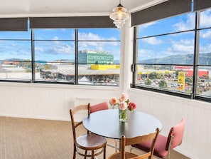 A room with a round table, chairs, a vase with flowers, and large windows offering a view of buildings and a parking lot.