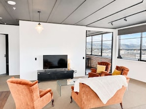 A modern living room with a flat-screen TV, leather sofas, a glass coffee table, and a large window offering a city view.