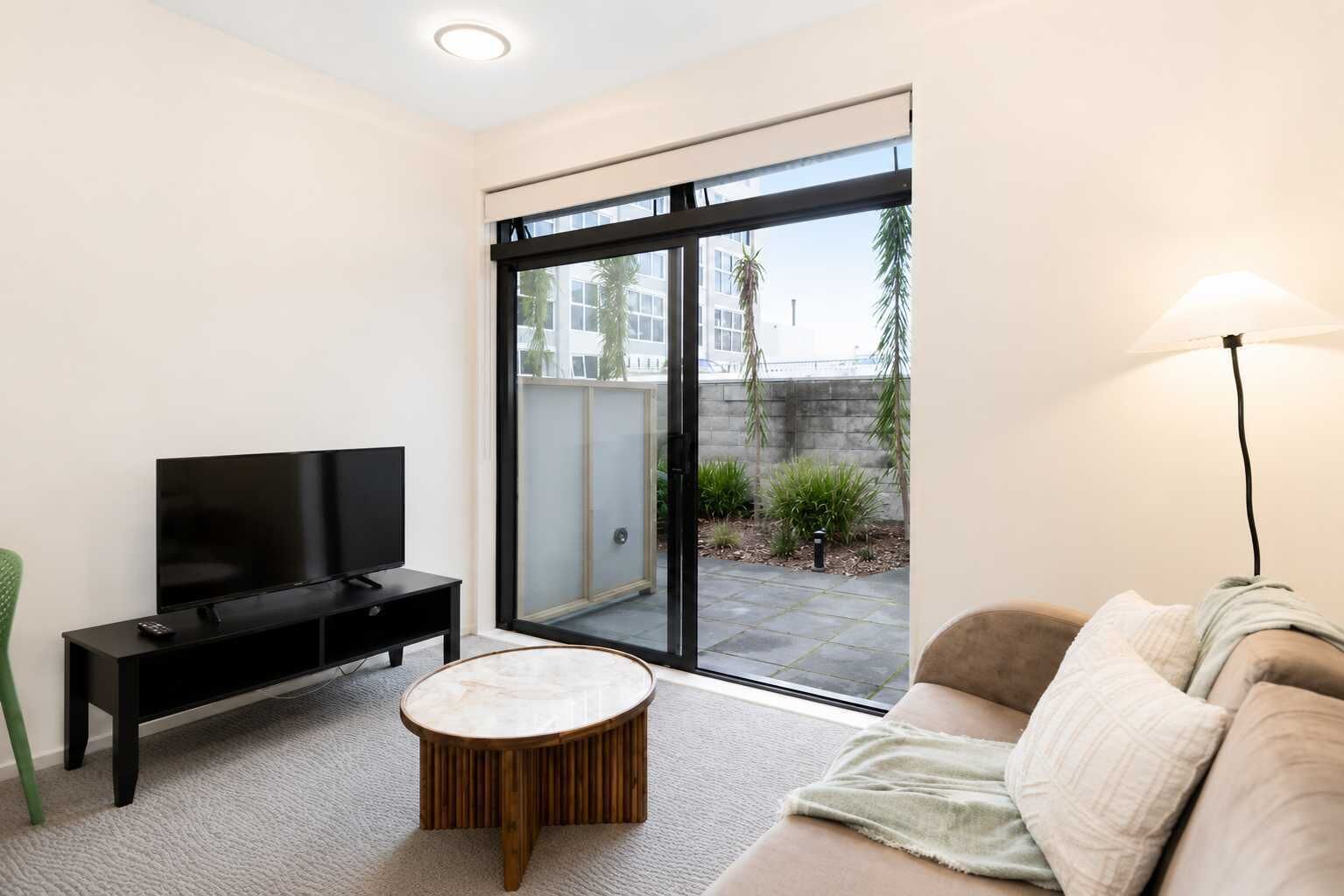 A modern living room with a flat-screen TV, a round coffee table, a sofa, and a sliding glass door leading to an outdoor area.