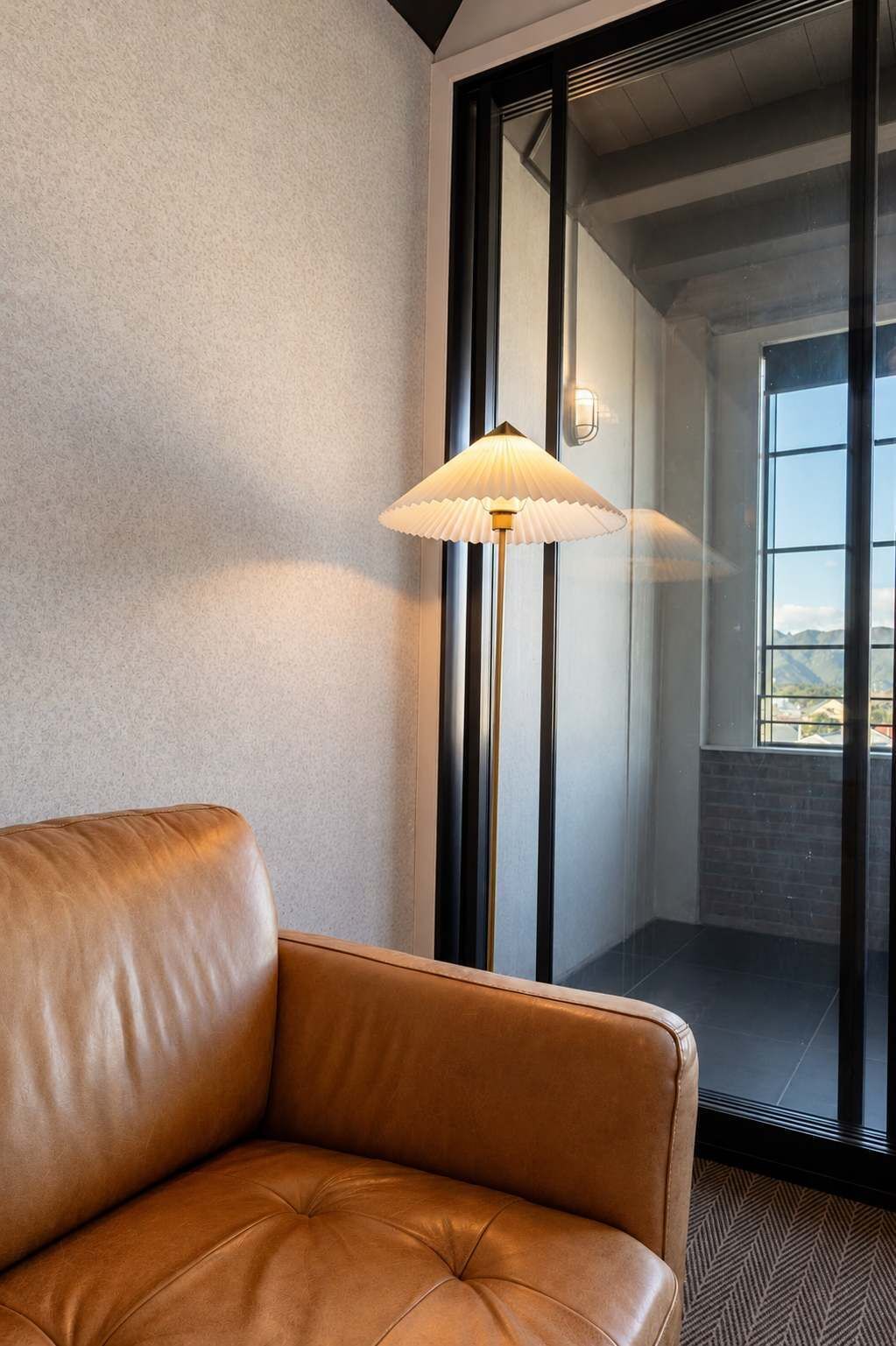 A leather sofa in a corner with a floor lamp and a view through a glass door to an interior space.