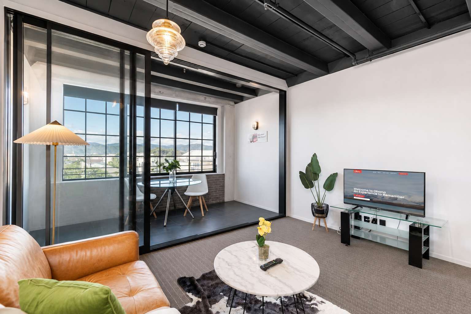 Uma sala de estar moderna com sofá de couro, mesa de centro redonda, televisão e uma janela ampla com vista para o exterior.