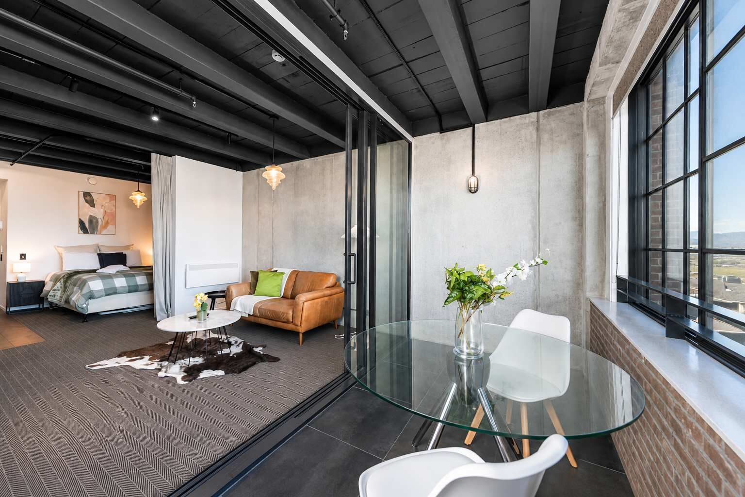 A modern hotel room with a glass dining table, a leather sofa, and a large window offering a city view.