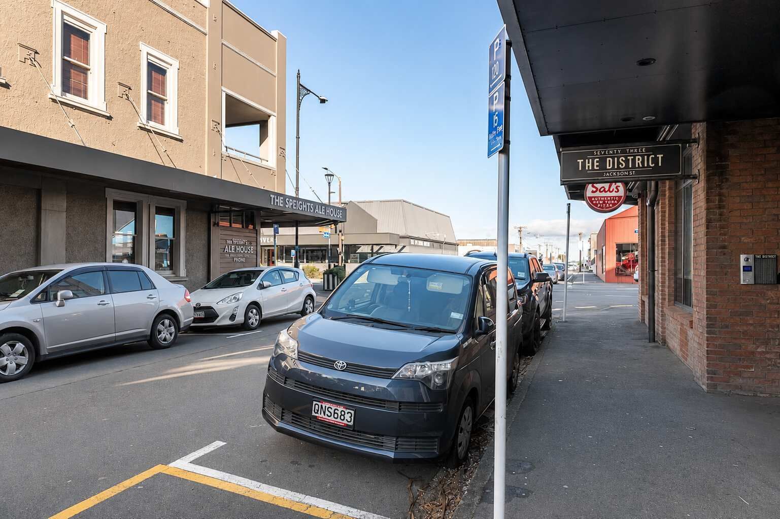 Vista de la calle con coches estacionados, un edificio con la leyenda 'The Speights Ale House' y un letrero que anuncia 'Sals Authentic Pizza'.