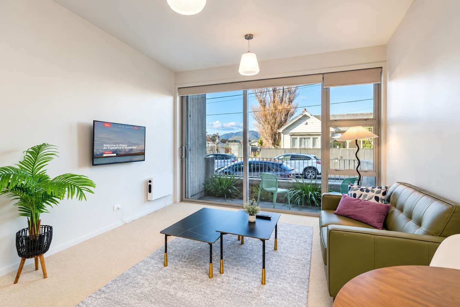 A modern living room with a flat-screen TV, a coffee table, a sofa, and a large window offering a view of the street and parked cars.