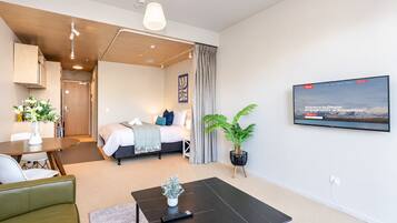 A modern hotel room with a bed, a dining area, a TV, and a potted plant.