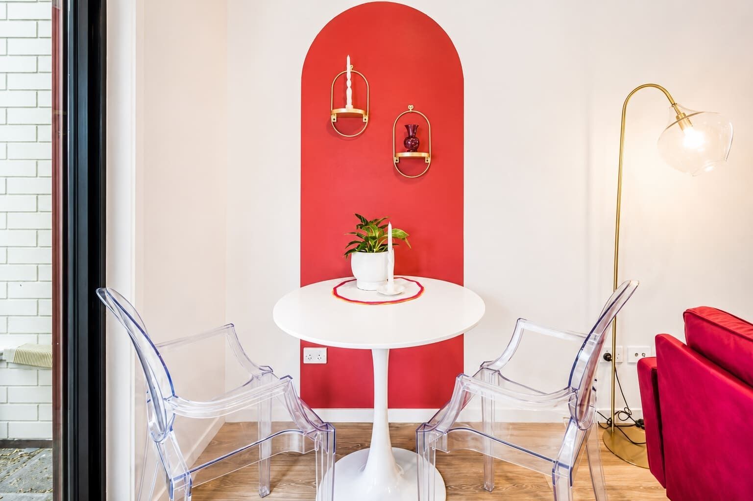 Una habitación con una mesa redonda blanca, dos sillas transparentes, una pared de acento roja, una planta en maceta y una lámpara de pie.