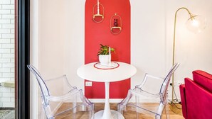 Una habitación con una mesa redonda blanca, dos sillas transparentes, una pared de acento roja, una planta en maceta y una lámpara de pie.