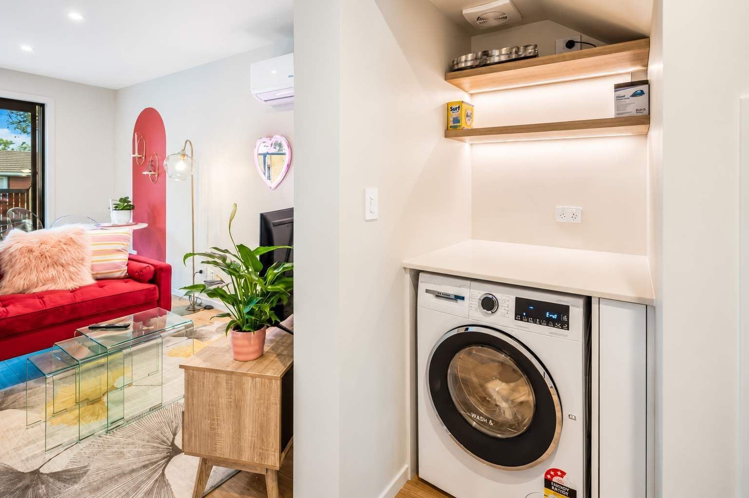 A compact living space with a red sofa, a glass coffee table, a potted plant, a washing machine, and a small wooden cabinet.