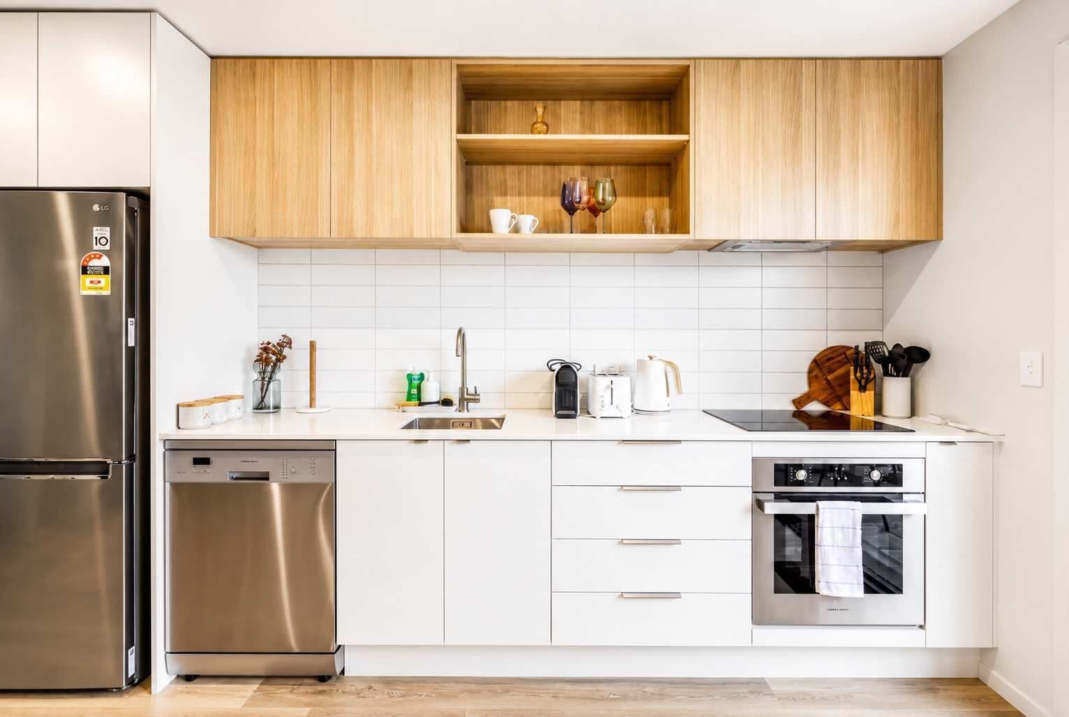 A modern kitchen with white cabinets, a stainless steel refrigerator, a dishwasher, a built-in oven, and a wooden shelf above the countertop.