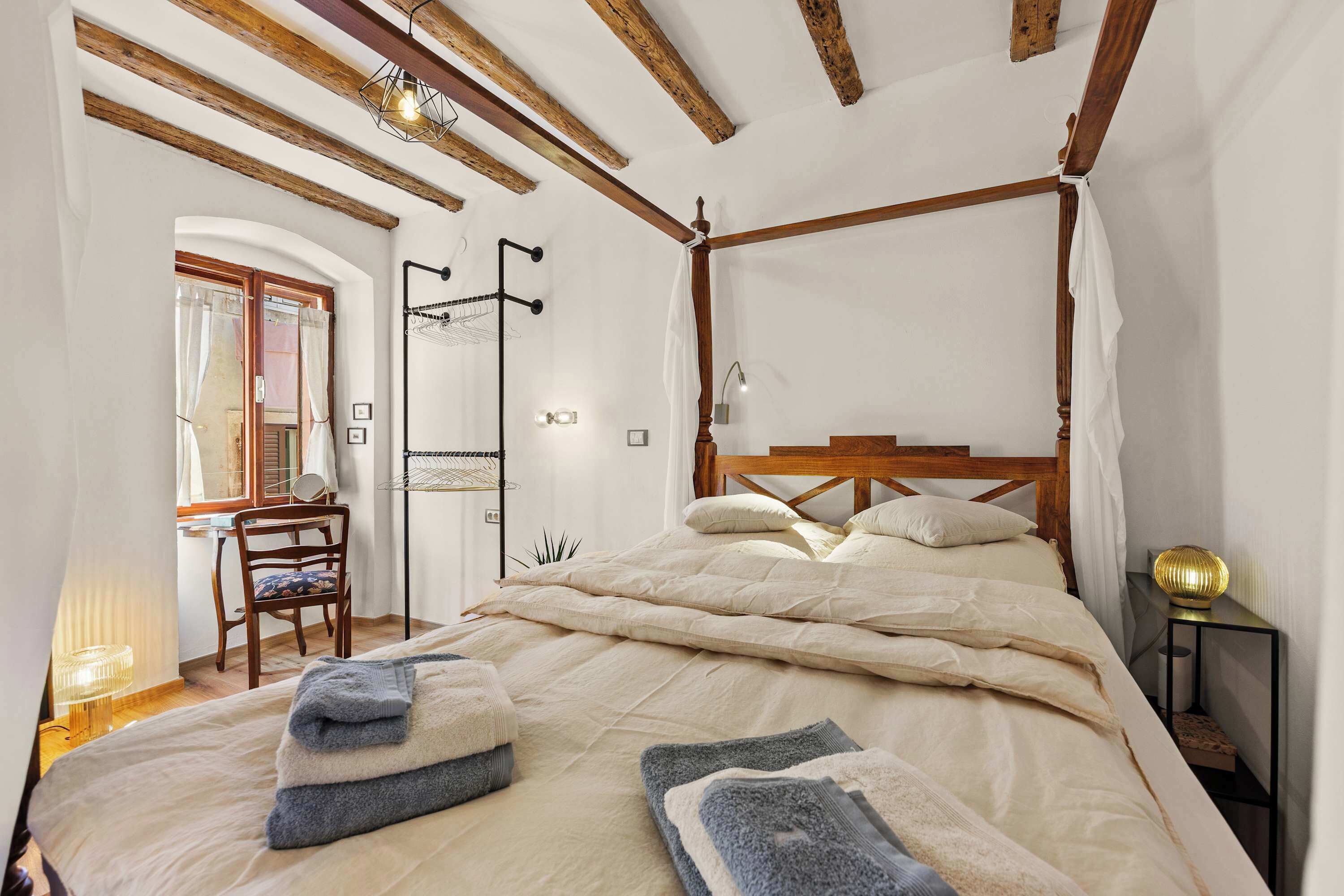 A bedroom with a four-poster bed, a wooden chair, a bedside table with a lamp, and a window with curtains.