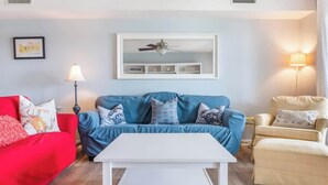 Ein Wohnzimmer mit blauem Sofa, rotem Sofa, weißem Couchtisch und beigem Sessel. Es gibt einen Deckenventilator, eine Wandleuchte und ein gerahmtes Bild an der Wand.