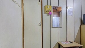 Una habitación con una puerta, una mesita con un control remoto, un tablón de anuncios y una rejilla metálica.