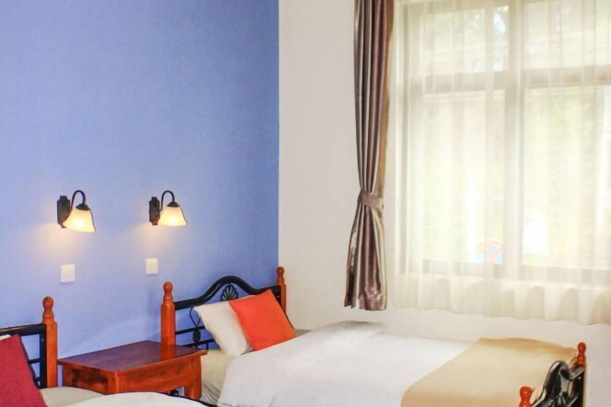 Una habitación de hotel con cama, una mesita pequeña, dos lámparas de pared y un ventanal con cortinas.