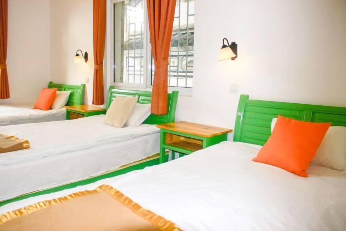 Habitación de hotel con dos camas individuales, cada una con cabecero verde y ropa de cama blanca. Hay dos mesitas de noche con lámparas y la habitación tiene una ventana con cortinas.