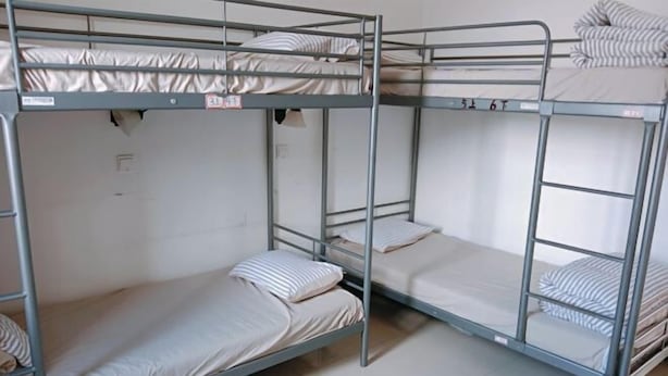 Habitación con literas de metal, ropa de cama blanca y un enchufe eléctrico visible.