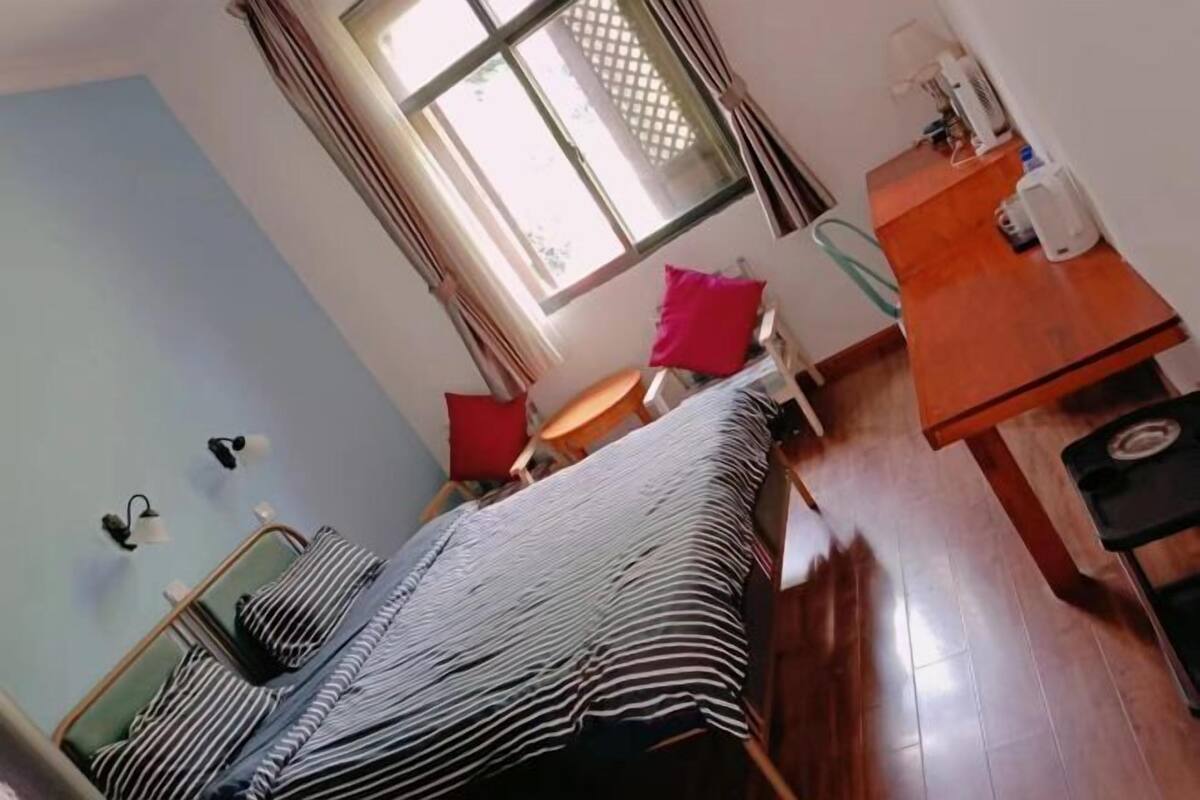 Una habitación con una cama, un escritorio, una silla, un ventanal con cortinas y una mesita con una botella.