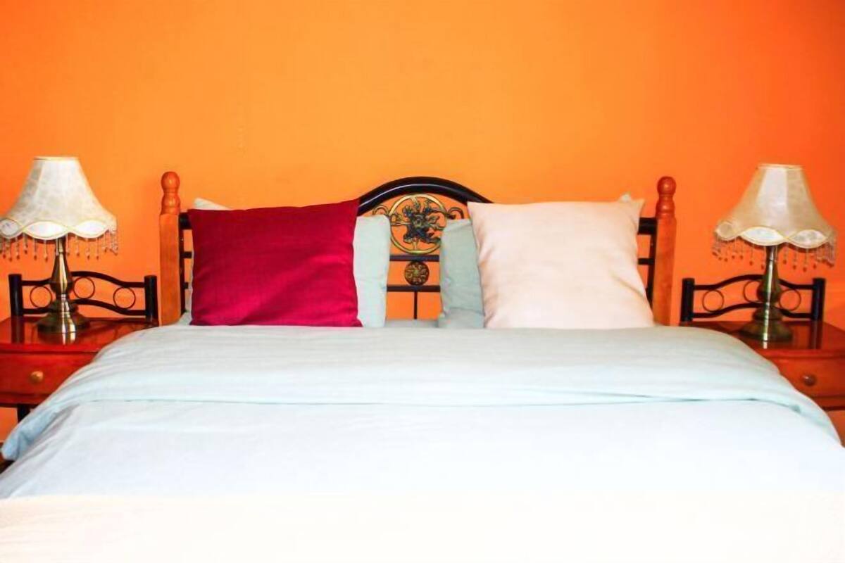 Una cama con cabecera de madera, dos lámparas de noche y un cojín rojo y blanco.