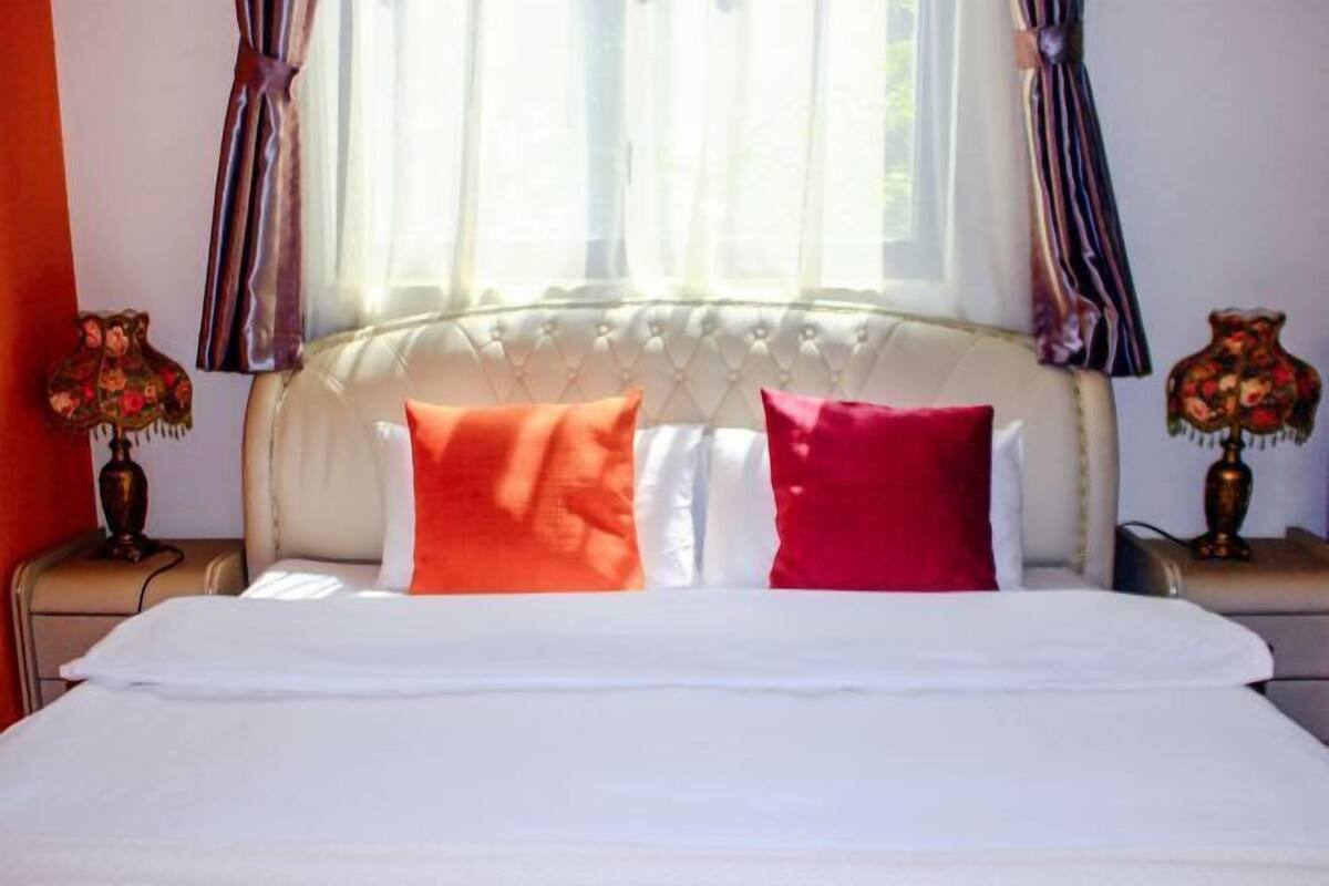 Una cama con almohadas blancas y rojas, un cabecero tapizado y cortinas.