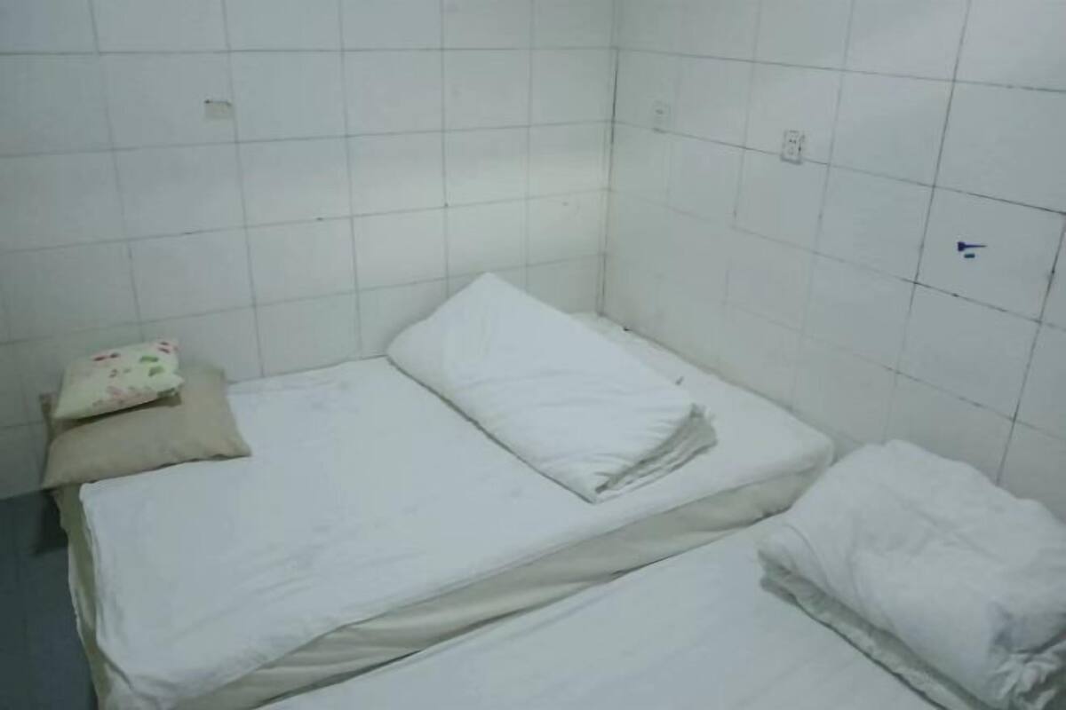 Una habitación pequeña con cama individual, paredes de azulejos blancos y una cama sencilla, sin adornos.