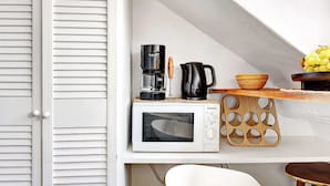 Een compacte keuken met een magnetron, koffiezetapparaat en een houten fruitschaal op een plank.