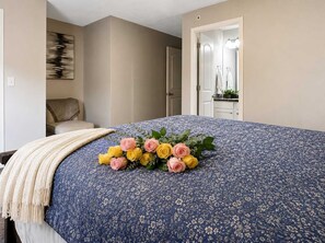 Un dormitorio con cama, mesita de noche, una silla, una puerta y un cuadro en la pared.