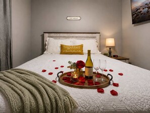 Una cama impecablemente arreglada con un cartel que dice 'Stay Awhile', un cojín con la frase 'Together our favorite place to be', una bandeja con pétalos de rosa, una botella y copas.