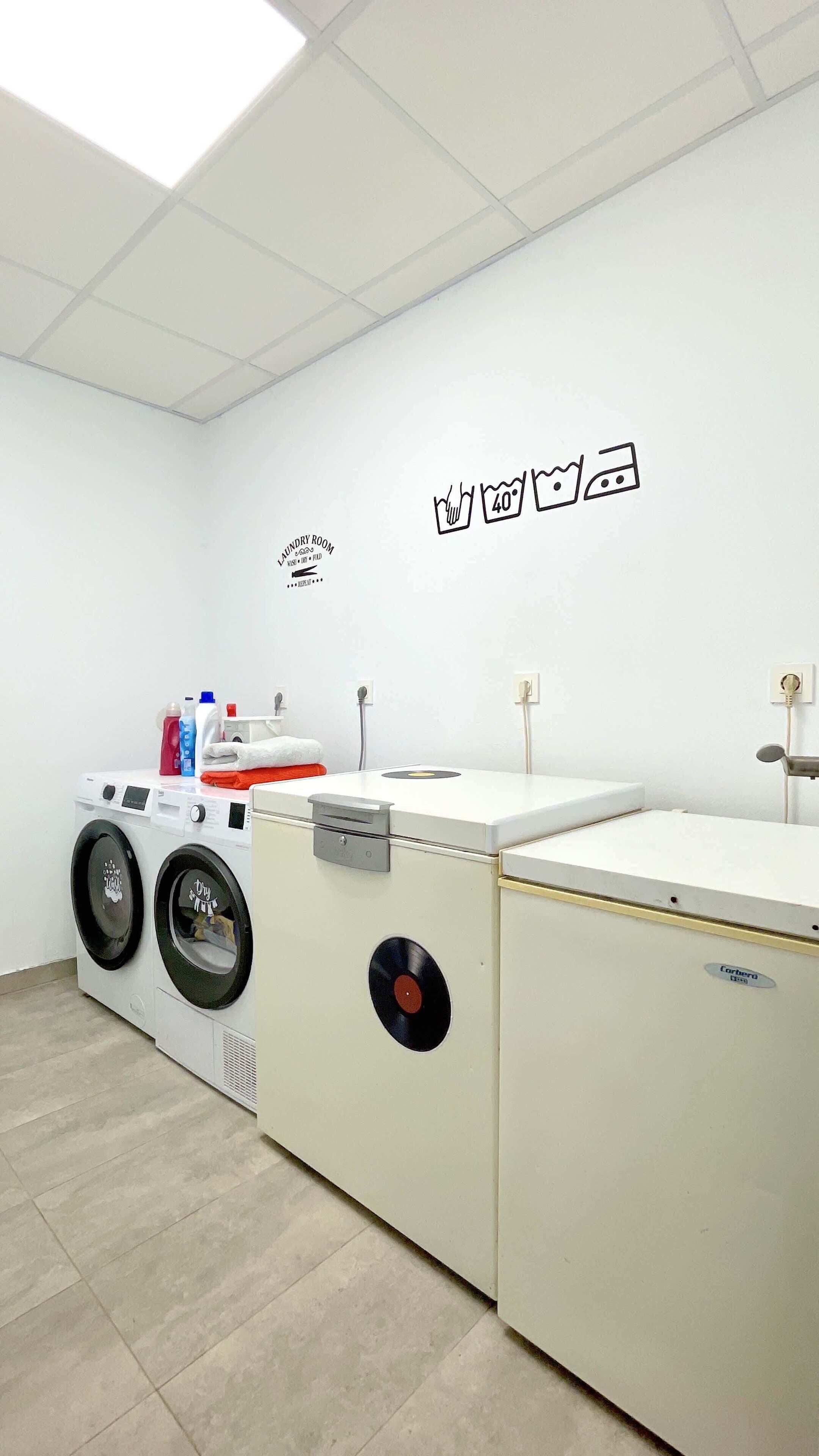 Een wasserette met twee wasmachines, een droger en een wandplank met wasbenodigdheden.