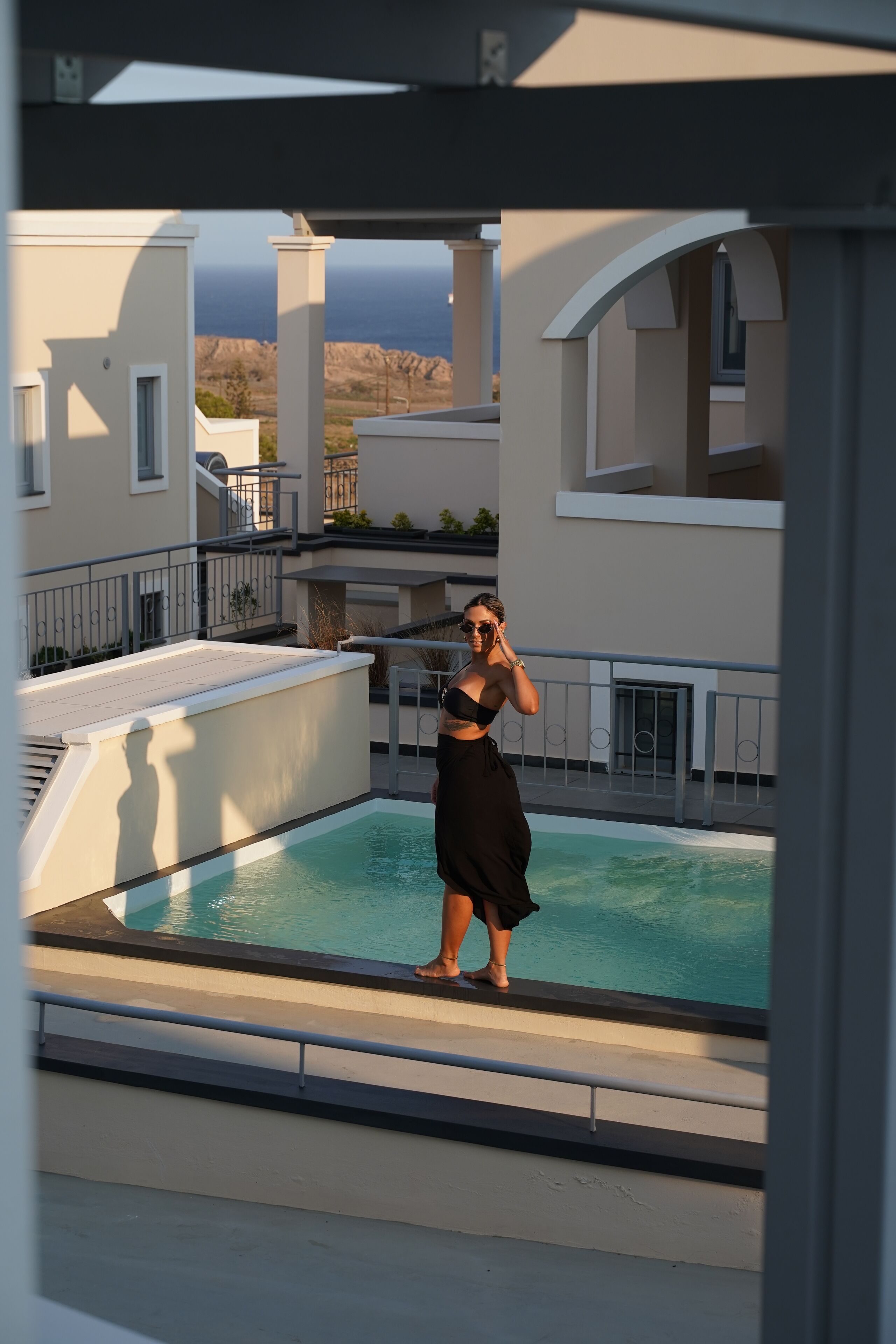 A person standing by a pool on a balcony with a view of the ocean and buildings in the background.