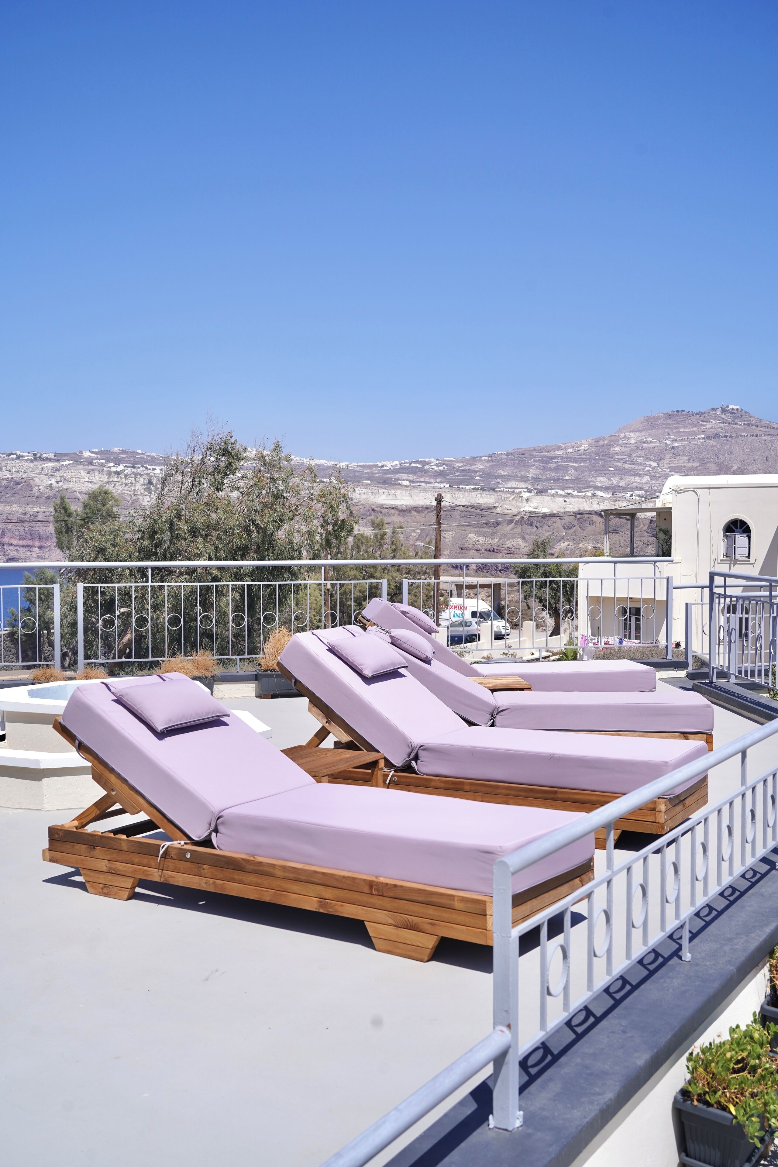 A rooftop terrace with four wooden sun loungers, a metal railing, and a view of a mountainous landscape.
