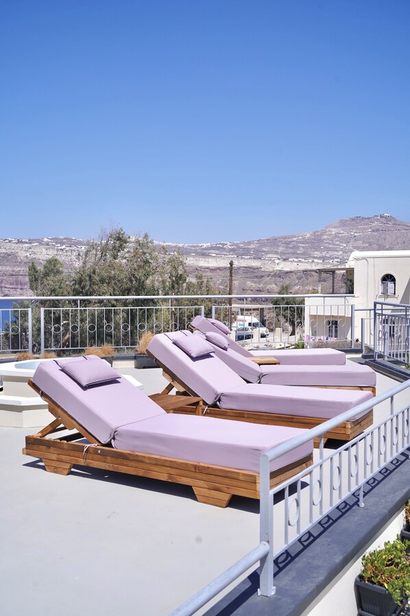 A rooftop terrace with four wooden sun loungers, a metal railing, and a view of a mountainous landscape.