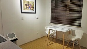 Ein Zimmer mit Holzboden, einem weißen Tisch mit Stühlen, einem gerahmten Bild an der Wand und einem Fenster mit Jalousien.