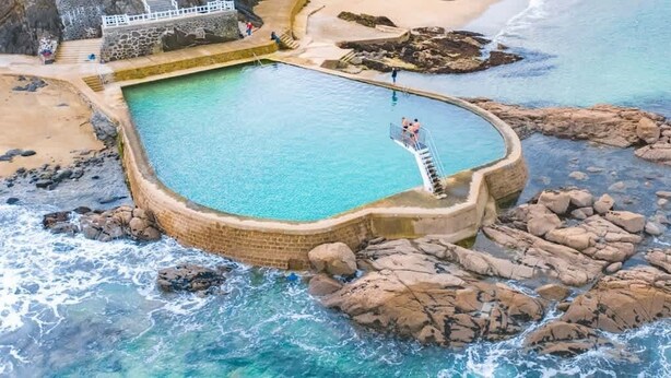An aerial view of a coastal swimming pool surrounded by rocky terrain and a beach.
